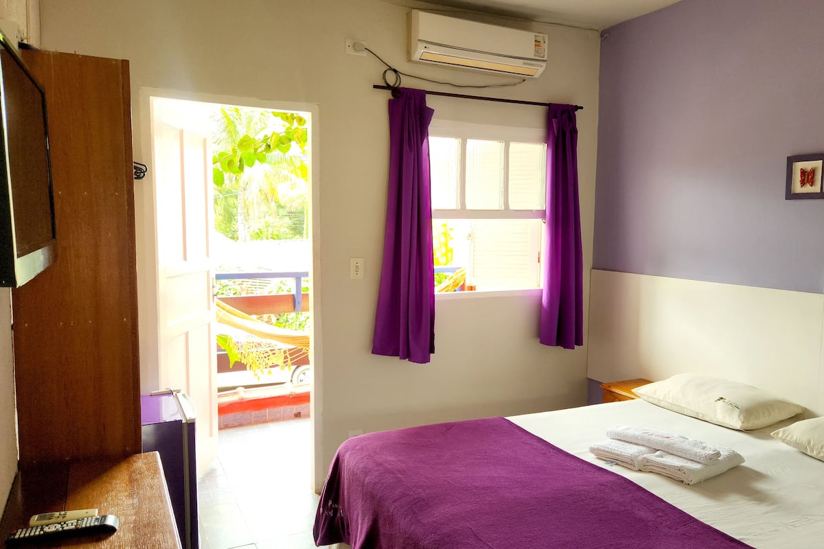 A bedroom with a bed, a window with purple curtains, an air conditioner, and a view of the outside.