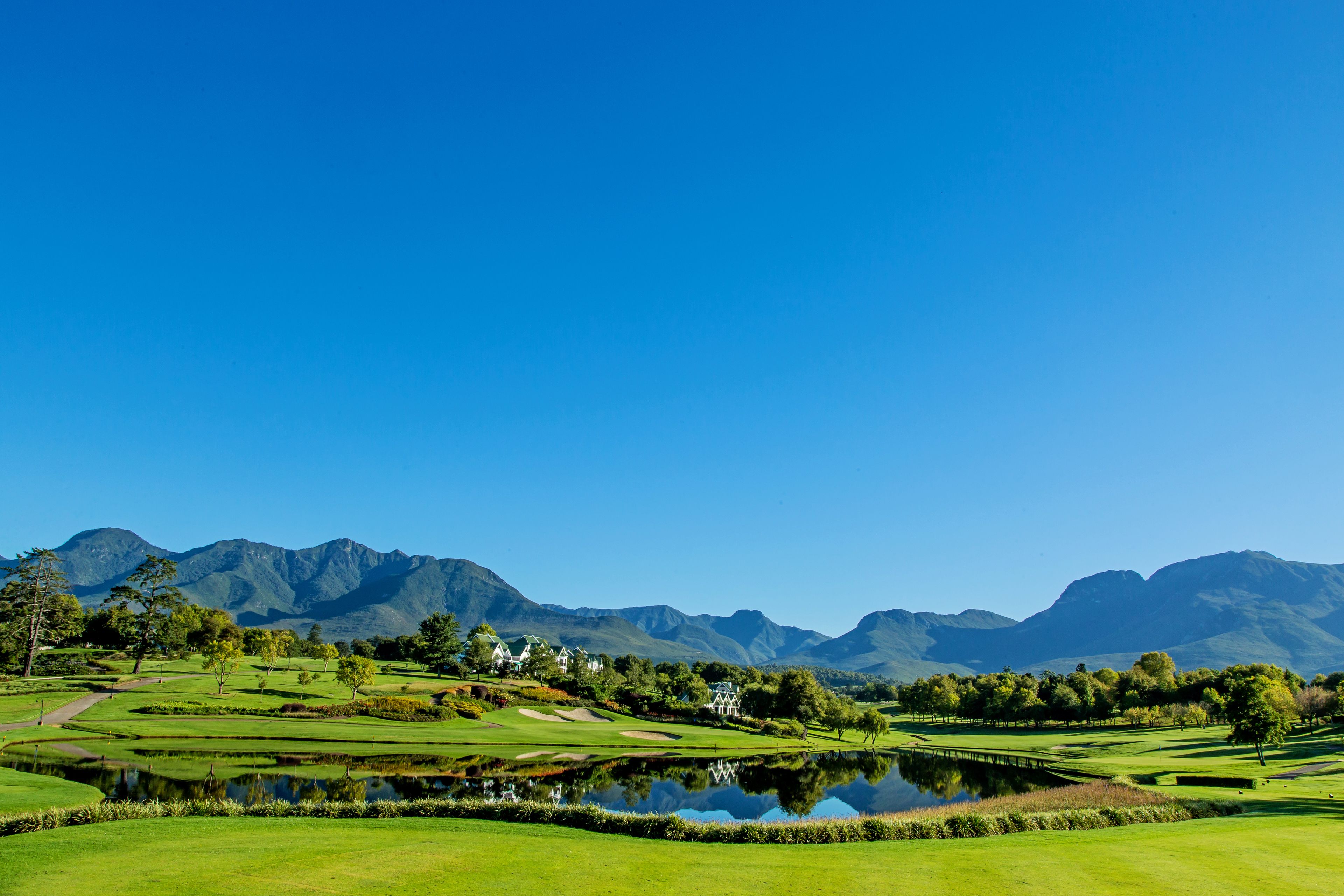 Un campo de golf con un estanque, casas y montañas al fondo.