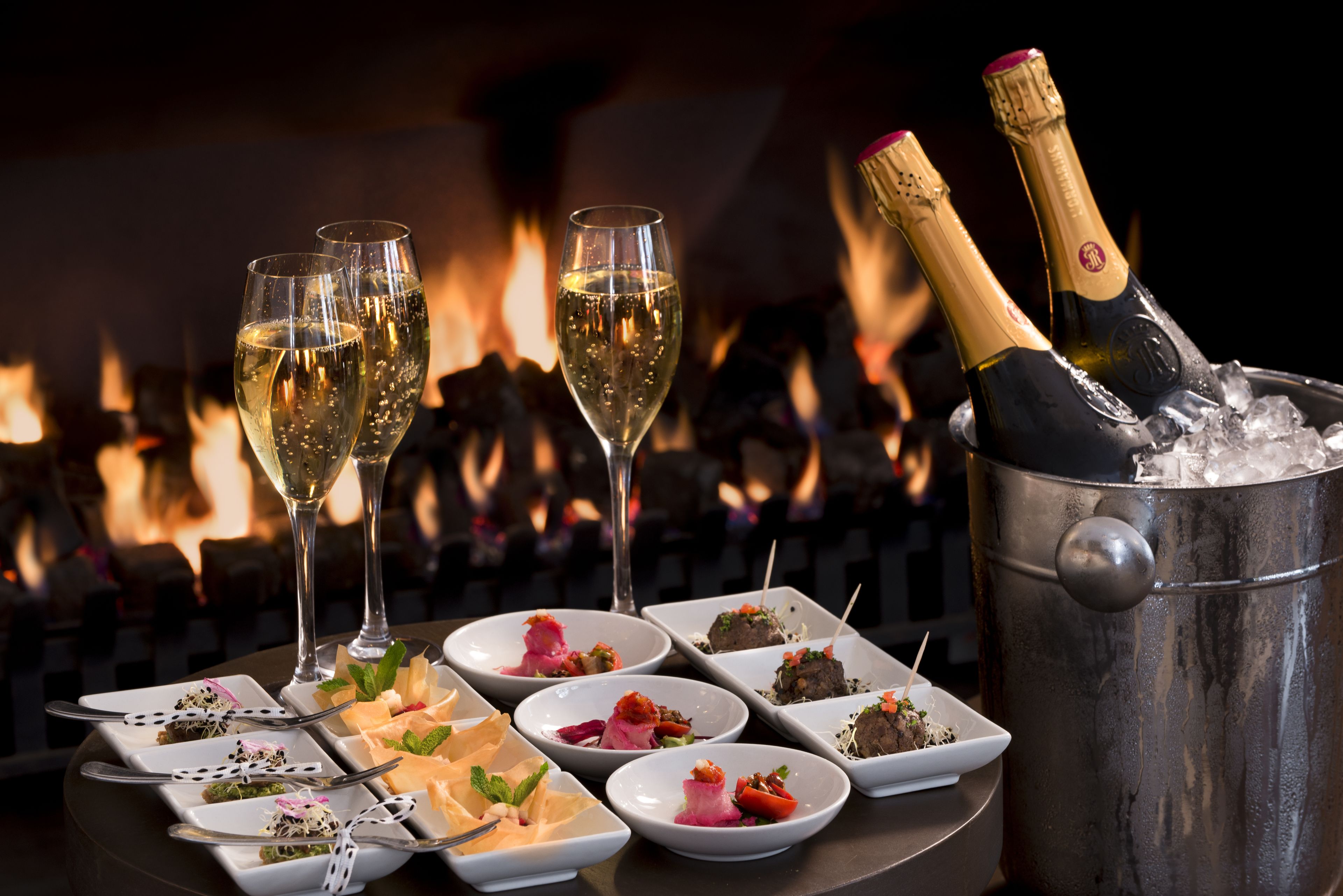 A table with multiple glasses of champagne and a bucket of ice, with a fire in the background.