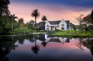 Una casa grande de estilo colonial blanco, con fachada simétrica, entrada central y un jardín bien cuidado con palmeras y un estanque.