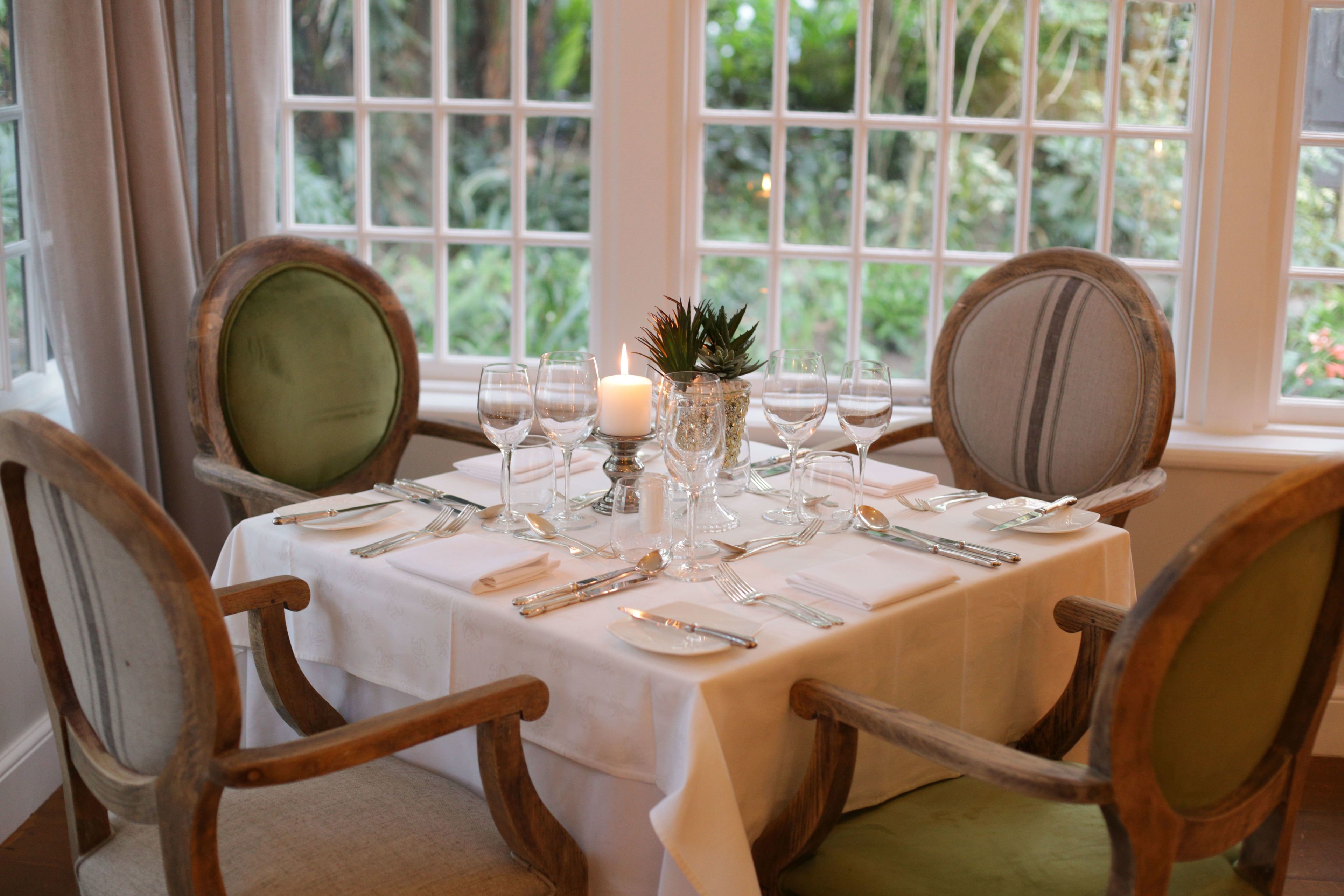 A dining table set for four with a white tablecloth, wooden chairs with green cushions, and a candle centerpiece.
