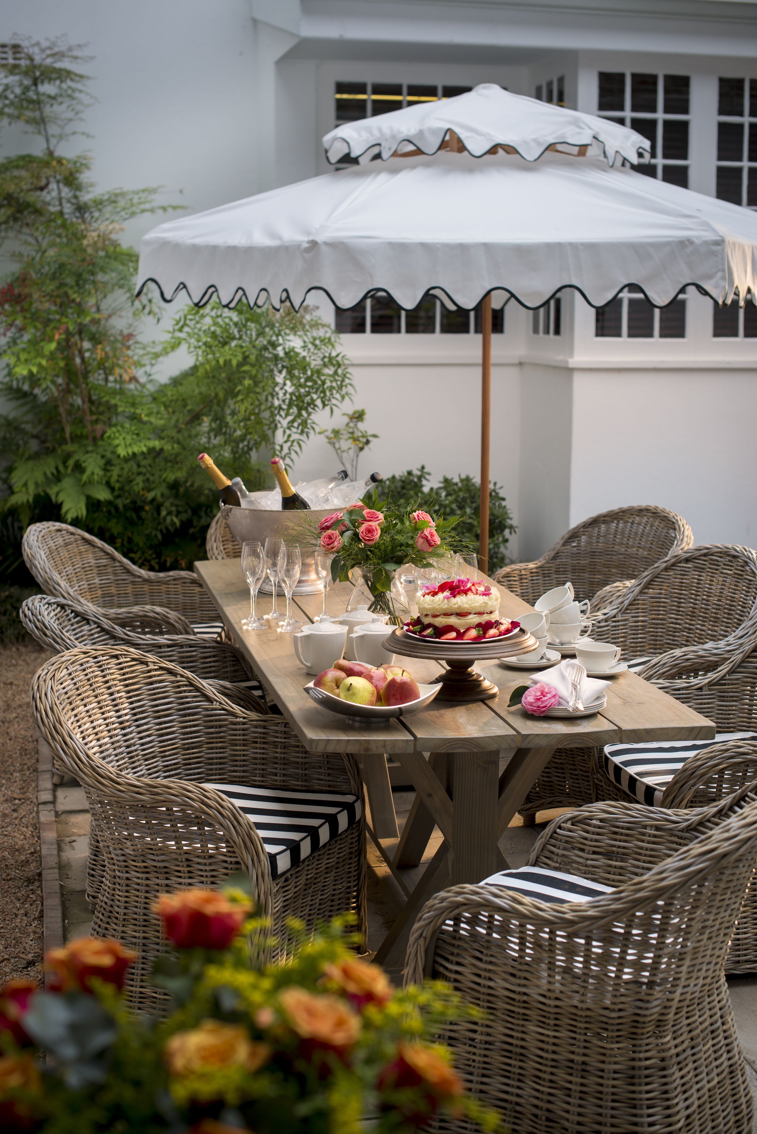 A dining table set outdoors with wicker chairs, a white umbrella, and a cake centerpiece.