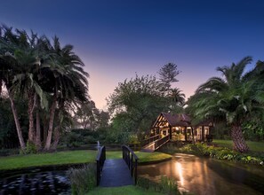 A wooden bridge leading to a house with a thatched roof, surrounded by palm trees and a pond. - The Manor House at Fancourt (George)