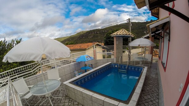 Ein Schwimmbecken mit gefliesten Rändern, umgeben von einer steinbelagten Terrasse und weißen Metallgeländern. Weiße Sonnenschirme und ein Tisch mit Stühlen sind vorhanden.