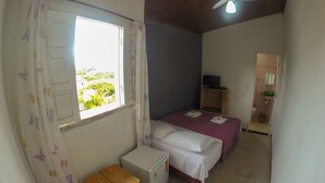 Ein kleiner, schlichter Raum mit einem Bett, einem kleinen Tisch, einem Kühlschrank, einem Deckenventilator und einem Fenster mit Vorhängen.