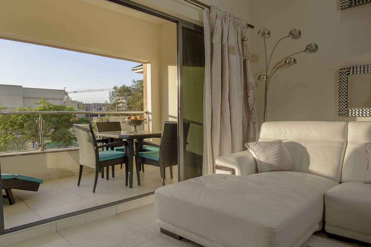 A balcony with a dining table and chairs, a sofa, and a large window offering a view of the cityscape.