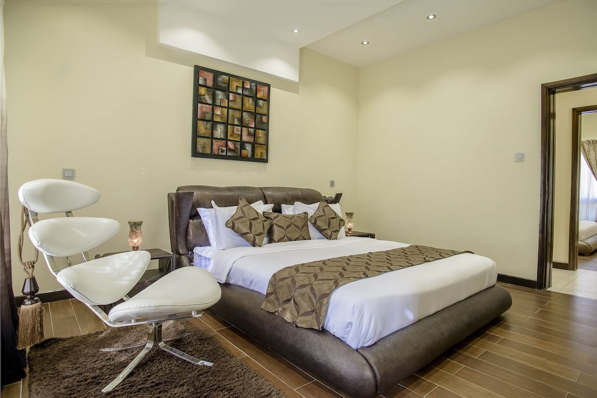 A modern bedroom with a large bed, a white chair, a decorative wall piece, and a wooden floor.