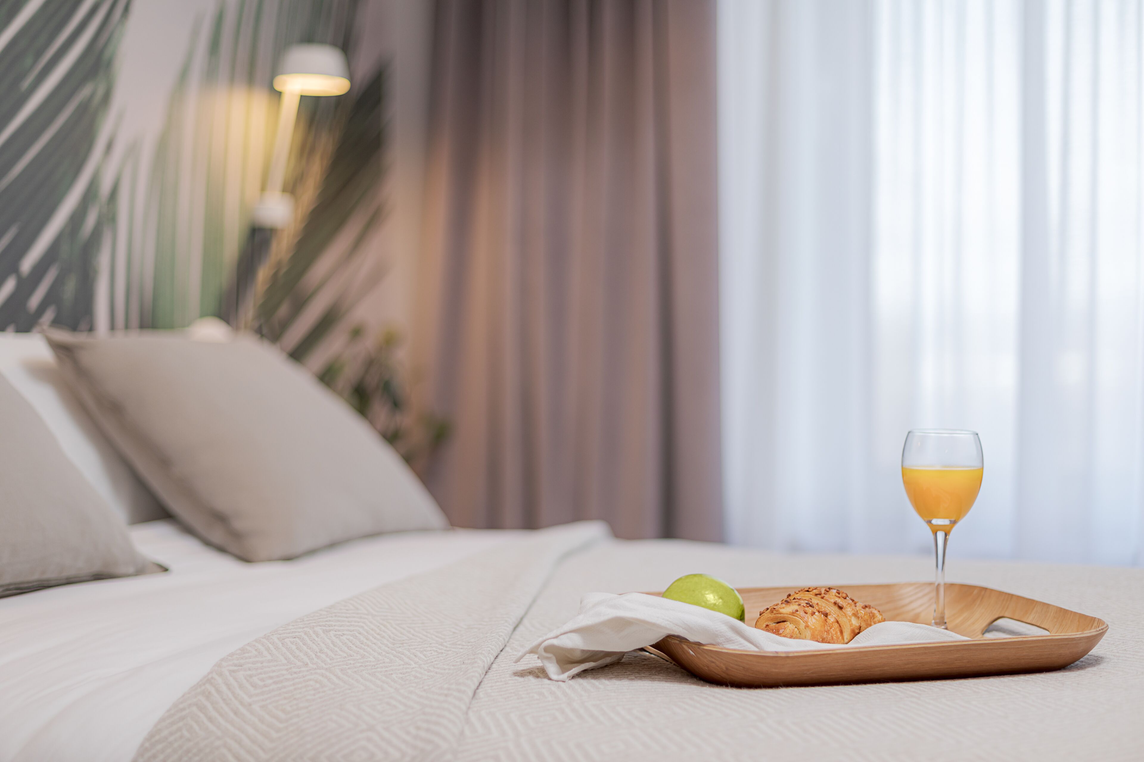 A neatly made bed with a breakfast tray containing a croissant, a glass of orange juice, and a green apple on a wooden tray.