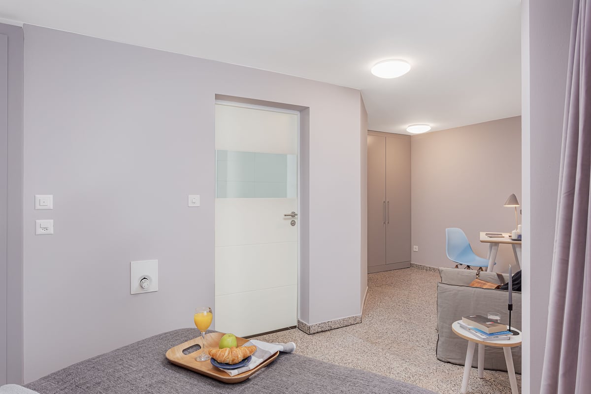 A modern hotel room with a grey carpet, a wooden tray with croissants and juice, a small table with a lamp, and a blue chair.