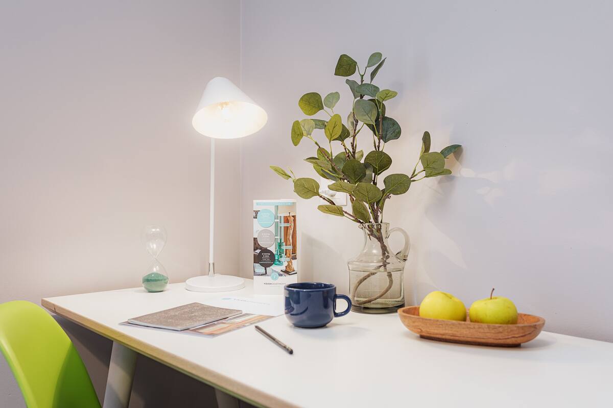A table with a lamp, a vase with green leaves, a blue mug, a bowl with apples, and a green chair.