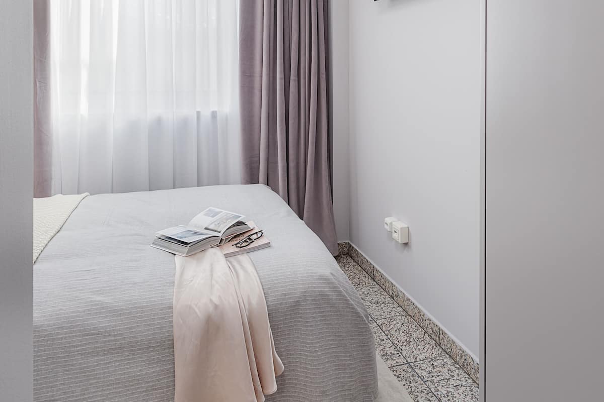 A bedroom with a bed, a flat-screen TV mounted on the wall, and a window with sheer curtains.