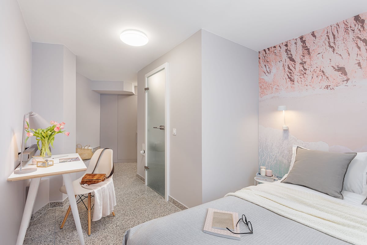 A hotel room with a bed, a desk with a chair, a small table with a vase of flowers, and a nightstand with a lamp.