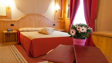Une chambre d’hôtel avec une tête de lit en bois, un lit recouvert d’une literie à motifs floraux, une table de chevet avec un téléphone, un vase rose contenant une fleur et une commode en bois.