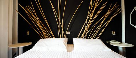 Una cama con una colcha blanca texturizada, dos almohadas blancas y un cabecero con listones de madera verticales. Hay dos mesitas de noche, una con tapa redonda de madera y la otra con superficie plana.