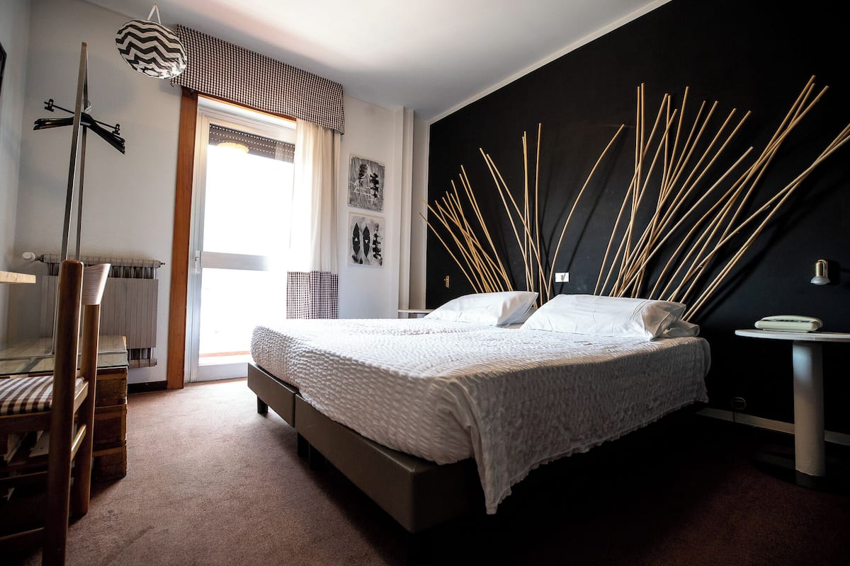 Un dormitorio con cama, una silla, un radiador, una ventana con cortinas y una pared decorada.