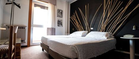 A bedroom with a bed, a chair, a radiator, a window with curtains, and a wall with decorative elements.
