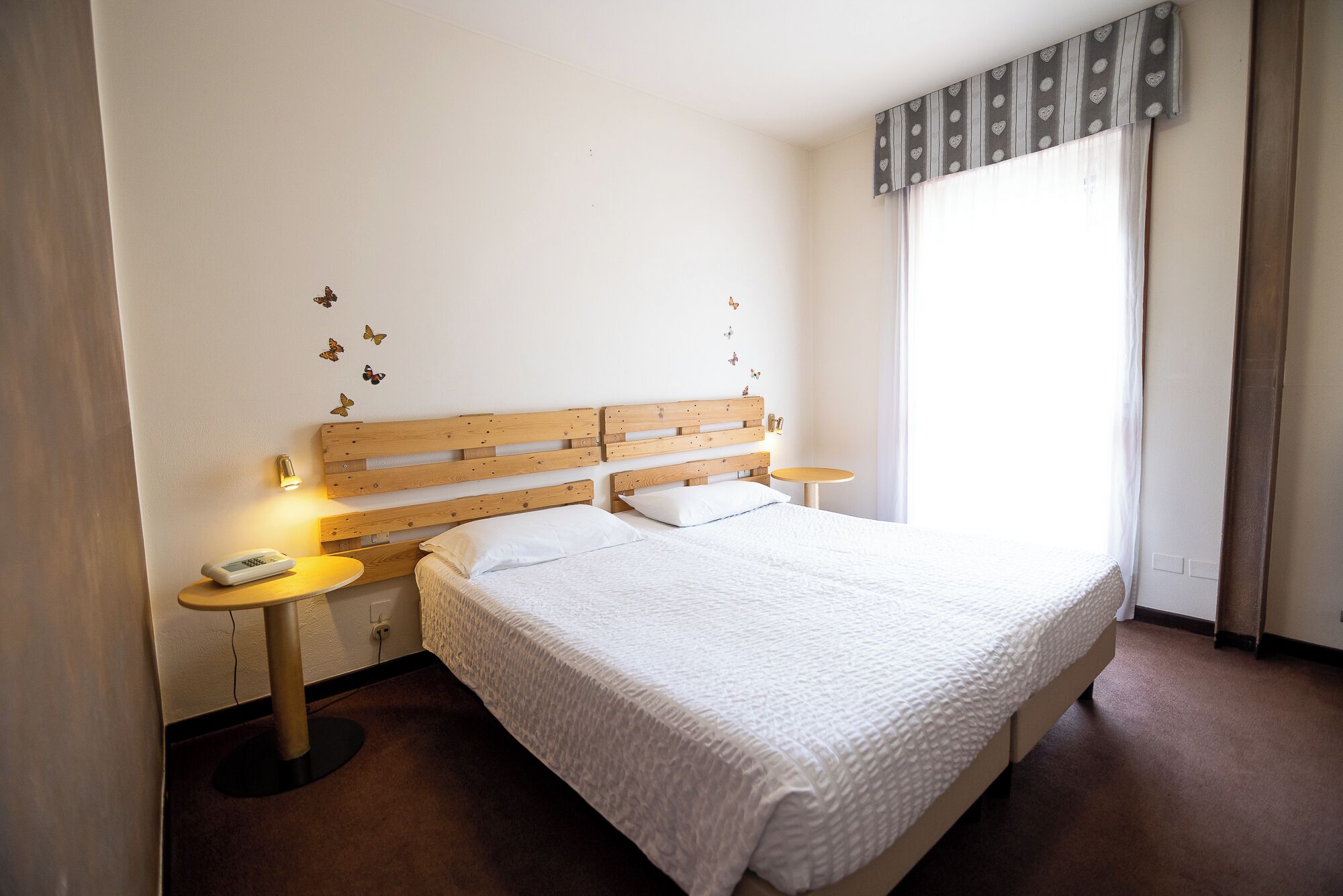 Un dormitorio con cabecera de madera, una cama individual, dos mesitas de noche, un teléfono en la pared y una ventana con cortinas.