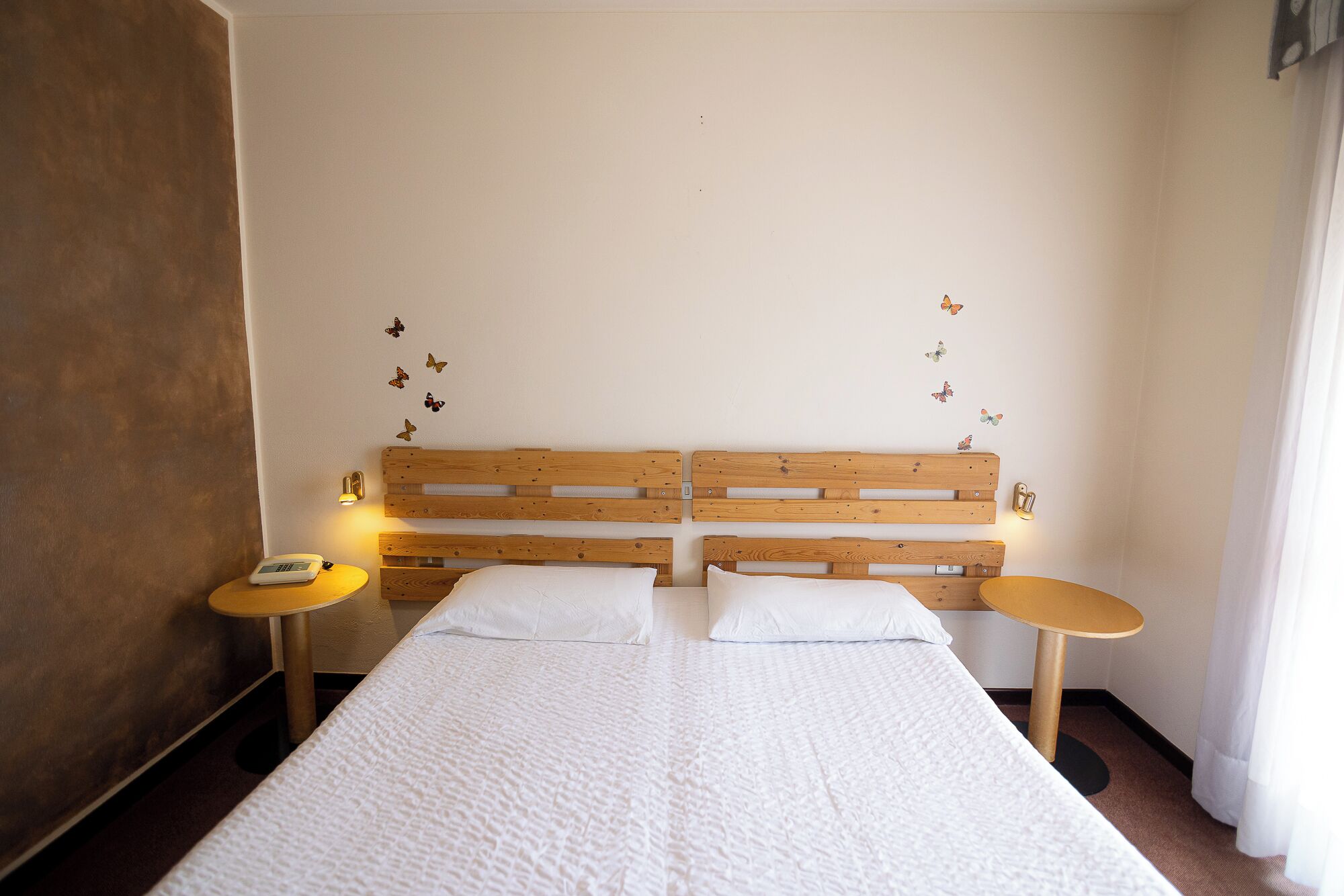 A room with a wooden headboard, two bedside tables with lamps, a bed with white bedding, and butterfly wall decals.