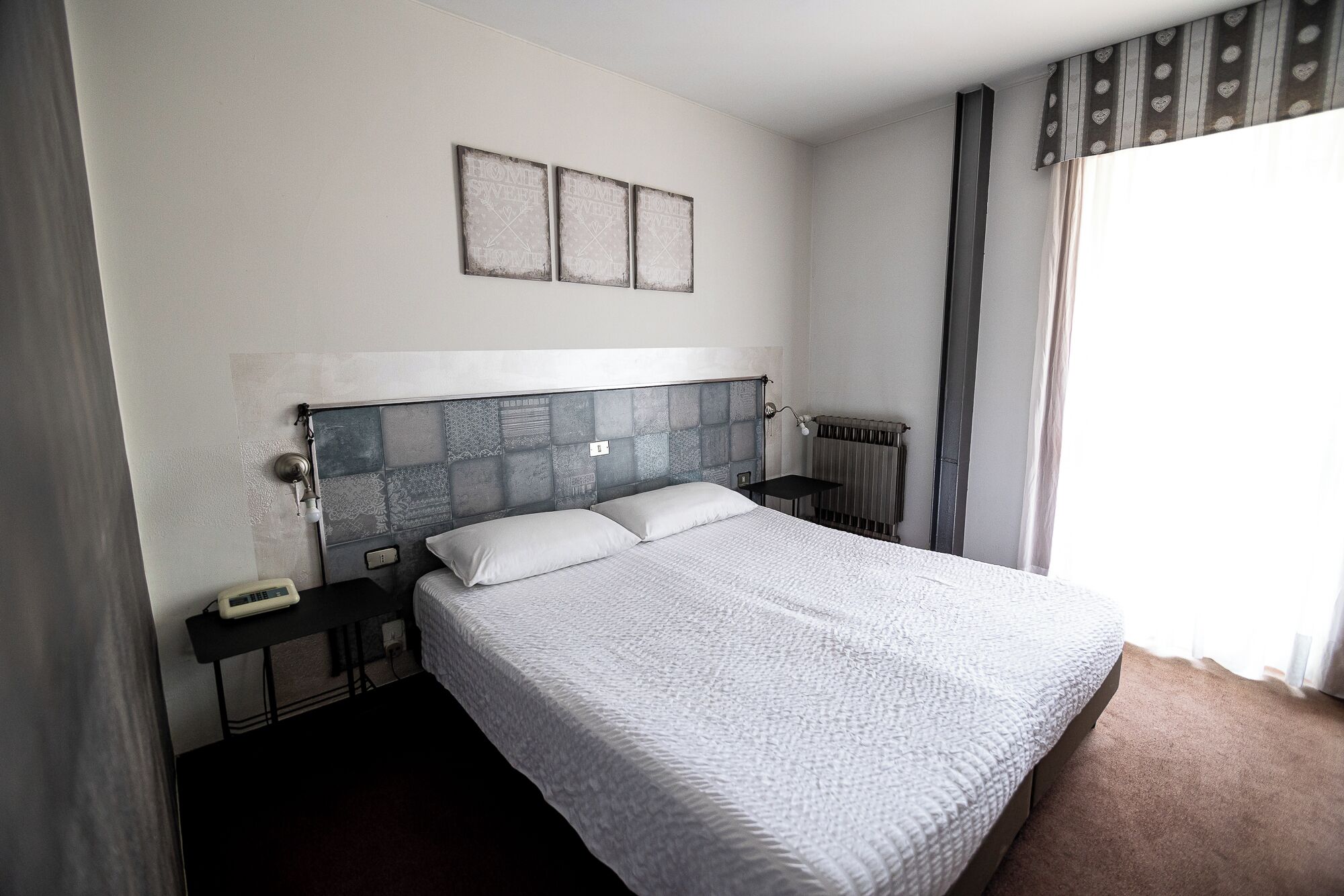 A hotel room with a large bed, a bedside table, a nightstand, a radiator, and two wall-mounted picture frames.
