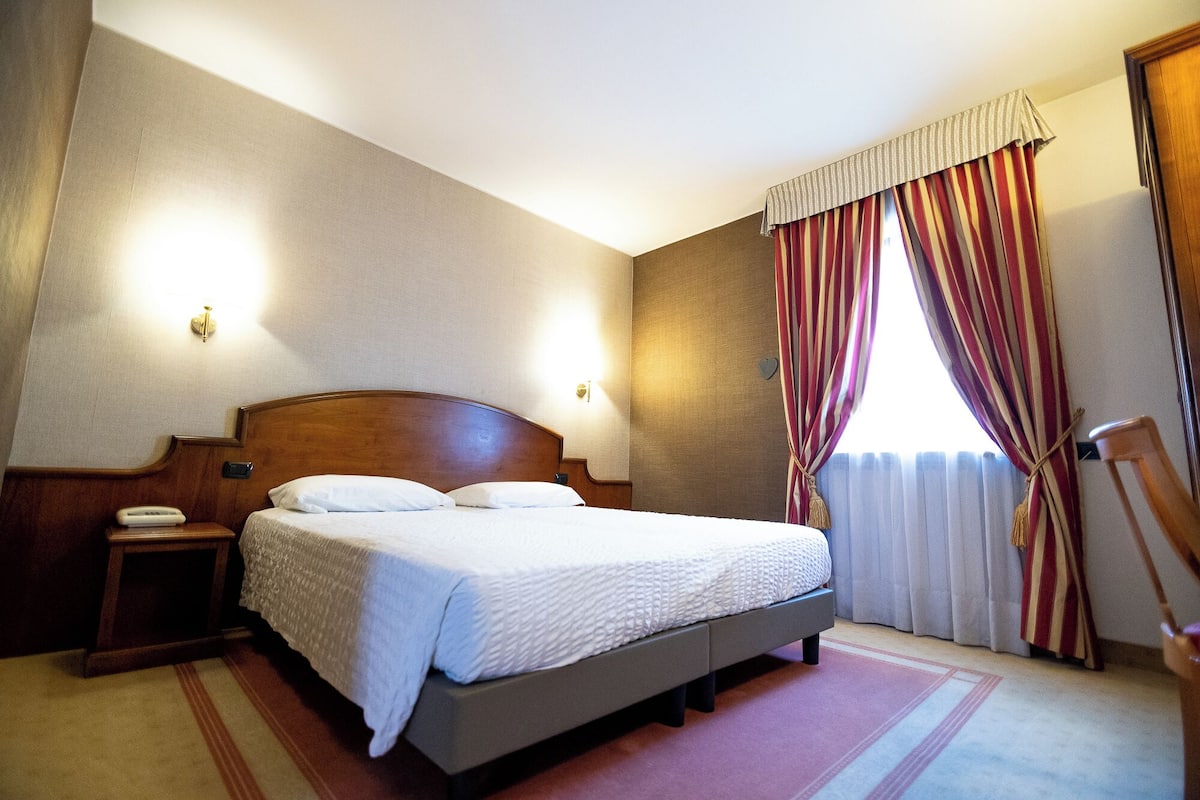 Una habitación de hotel con cama, mesitas de noche, una ventana con cortinas y una silla de madera.