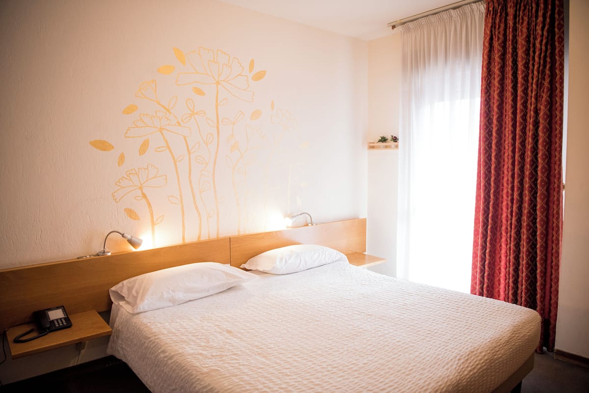 Una habitación de hotel con cama, mesita de noche, lámpara de pared y ventana con cortinas.