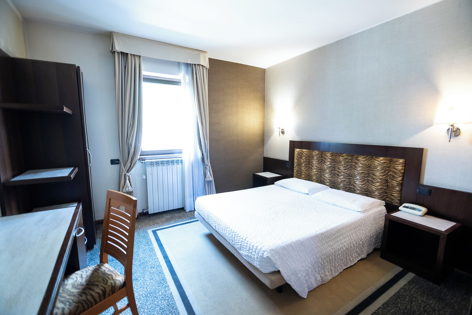 Une chambre d’hôtel comprenant un lit, un bureau, une chaise, une armoire et une fenêtre avec des rideaux.