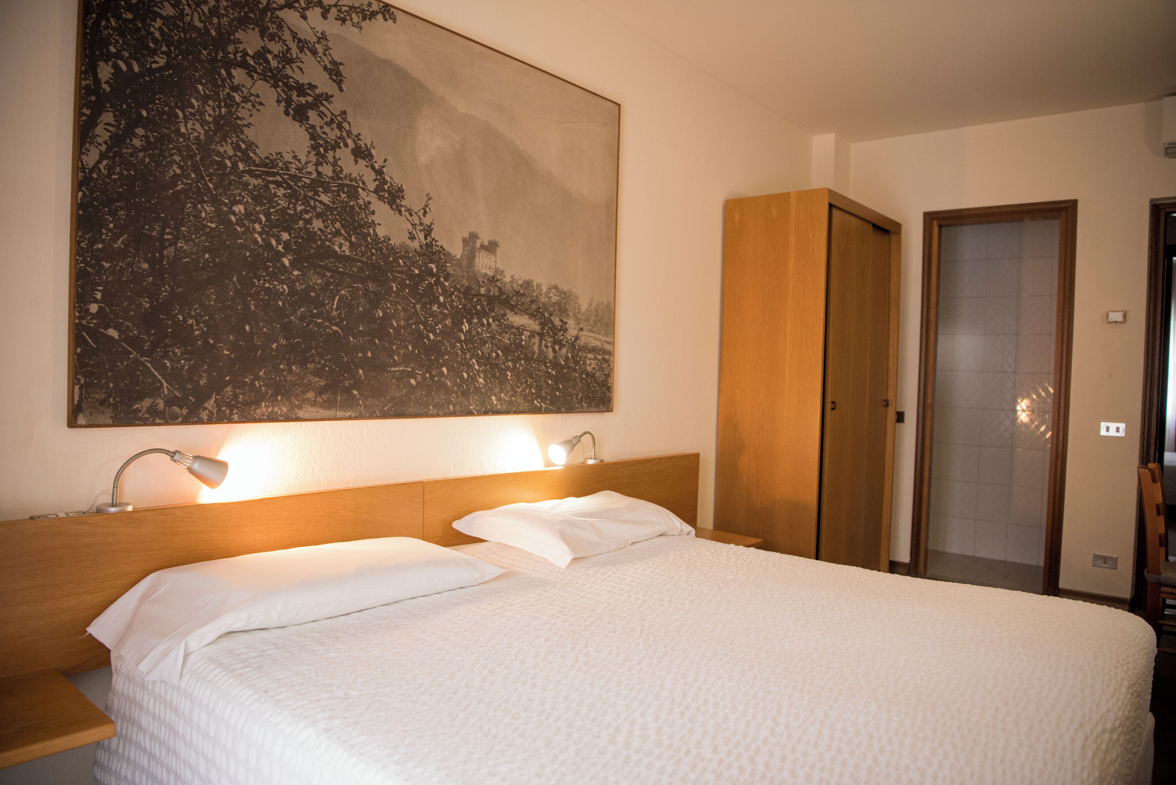 Een opgemaakt bed met witte lakens, een groot gepresenteerd schilderij aan de muur, en een houten garderobe.