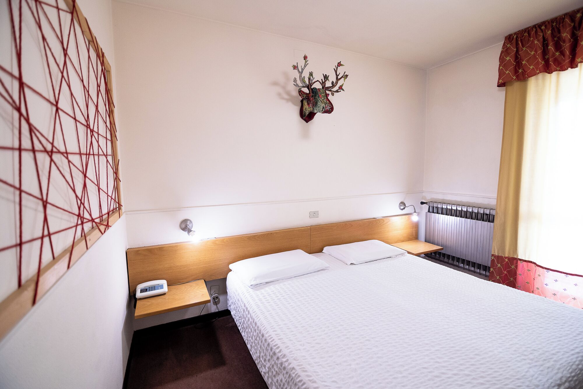 A hotel room with a bed, bedside tables, a wall-mounted phone, a radiator, and a decorative deer head on the wall.