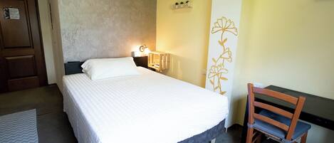 A hotel room with a bed, a desk, and a chair. There is a wall-mounted shelf with plants and a decorative wall piece.