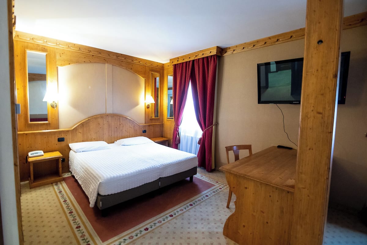Habitación de hotel con cama, mesita de noche, lámpara, espejo, televisor y escritorio de madera.
