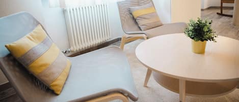 Una habitación con dos sillones tapizados grises con cojines amarillos y marrones, una mesa redonda de madera y una pequeña planta en maceta.