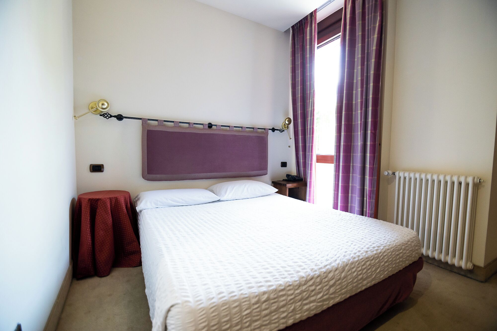 A bedroom with a bed, a nightstand, a red chair, a window with curtains, and a radiator.