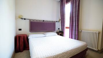 Une chambre à coucher avec un lit, une table de chevet, un fauteuil rouge, une fenêtre avec des rideaux et un radiateur.