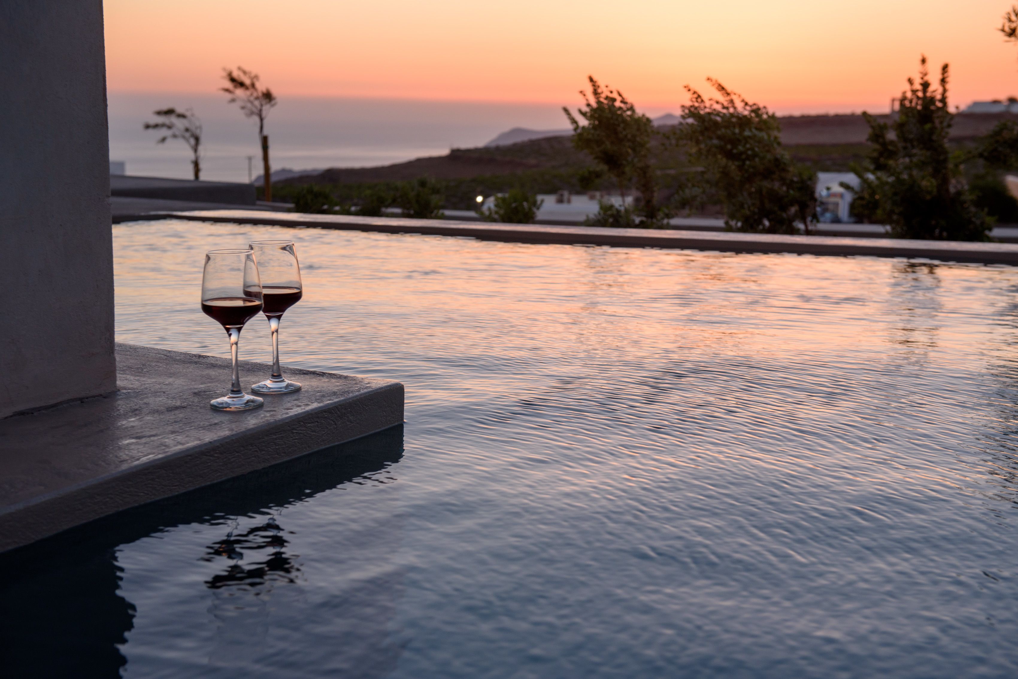 A poolside view with two glasses of red wine, calm water, and a sunset in the background.