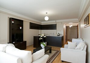 A modern living room with a sofa, a coffee table, and a TV. There is a kitchen area with a sink and a countertop.