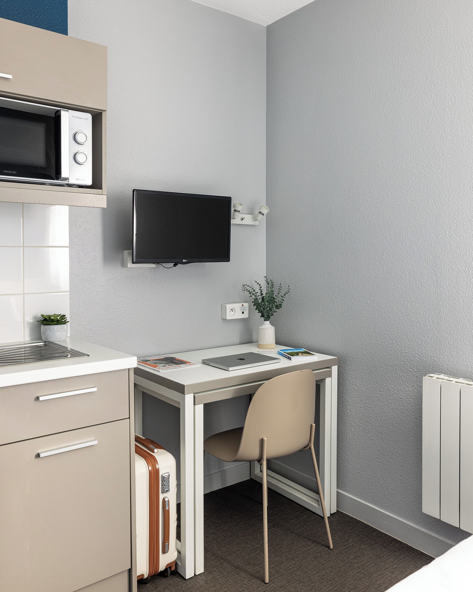 A modern kitchen with a microwave, a flat-screen TV mounted on the wall, a small potted plant, and a desk with a chair.