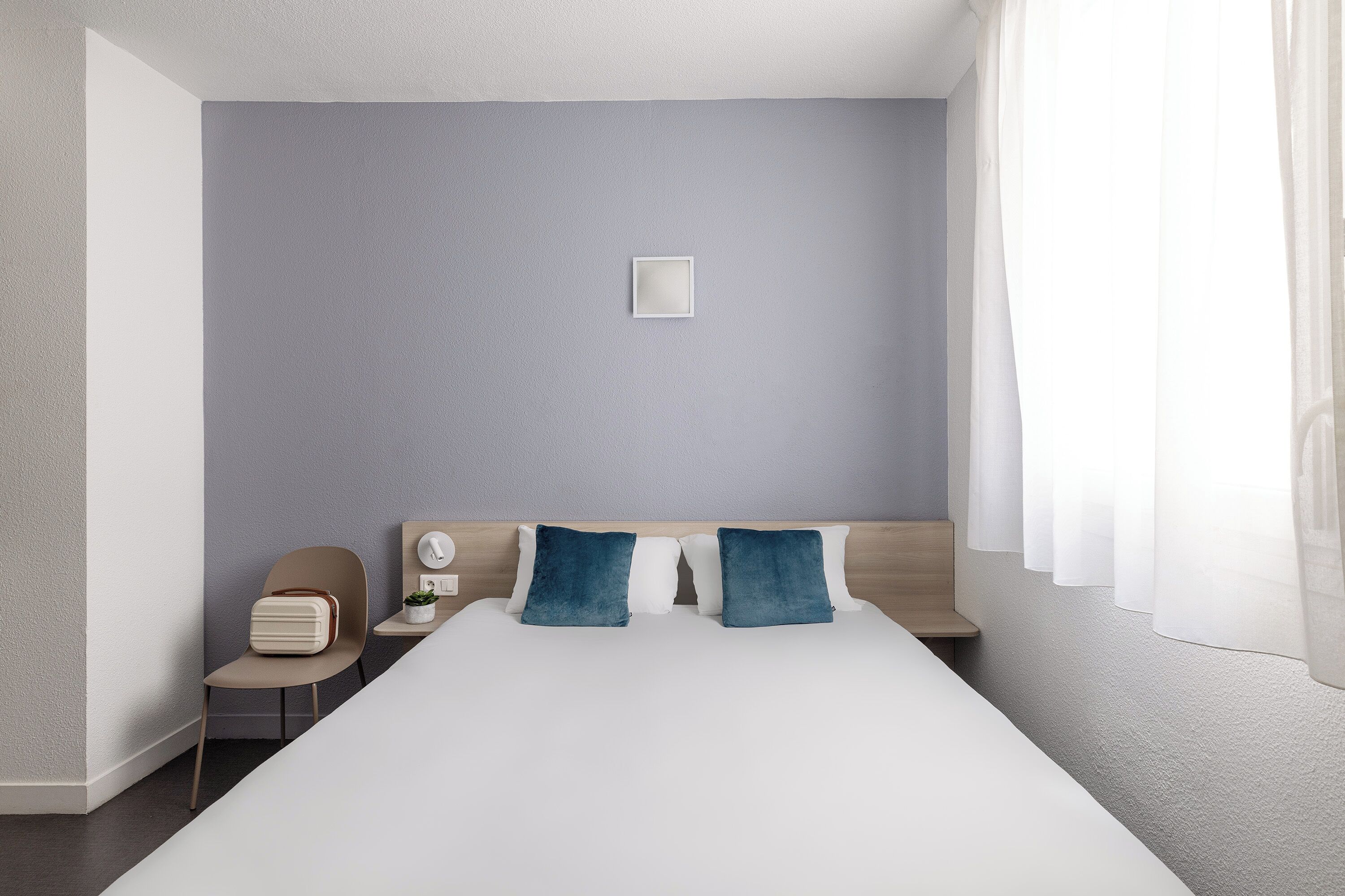 A modern hotel room with a large bed, a bedside table, a chair, and a window with curtains.