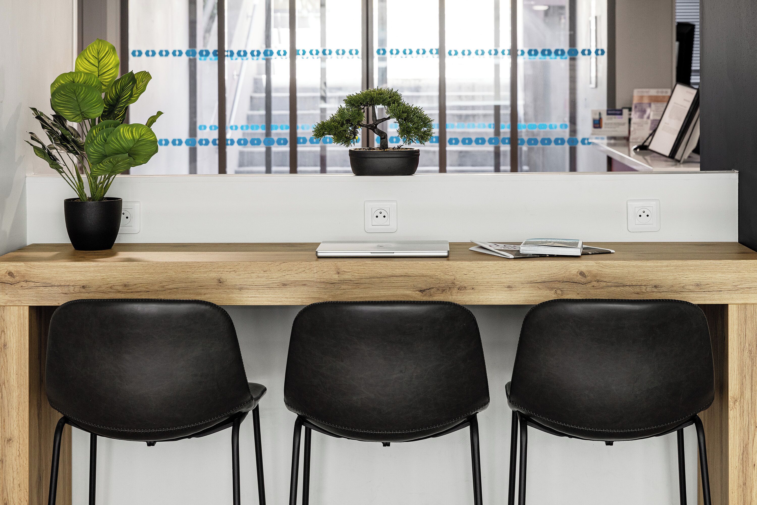 Un espace de travail moderne avec un bureau en bois, trois chaises noires, une plante en pot et une fenêtre avec des stores.