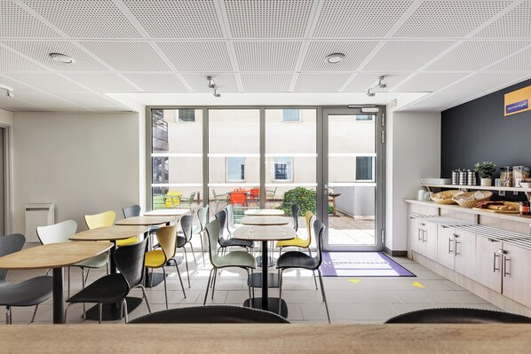 Un comedor moderno con mesas y sillas, una zona de cocina con armarios y una encimera, y ventanas amplias que dejan entrar luz natural.