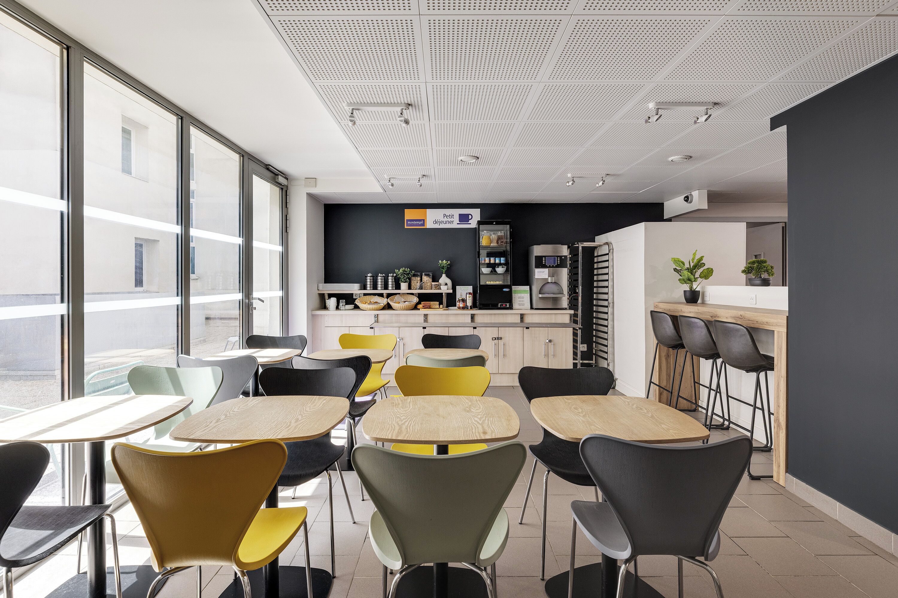 A modern dining area with tables and chairs, a kitchen in the background, and large windows.