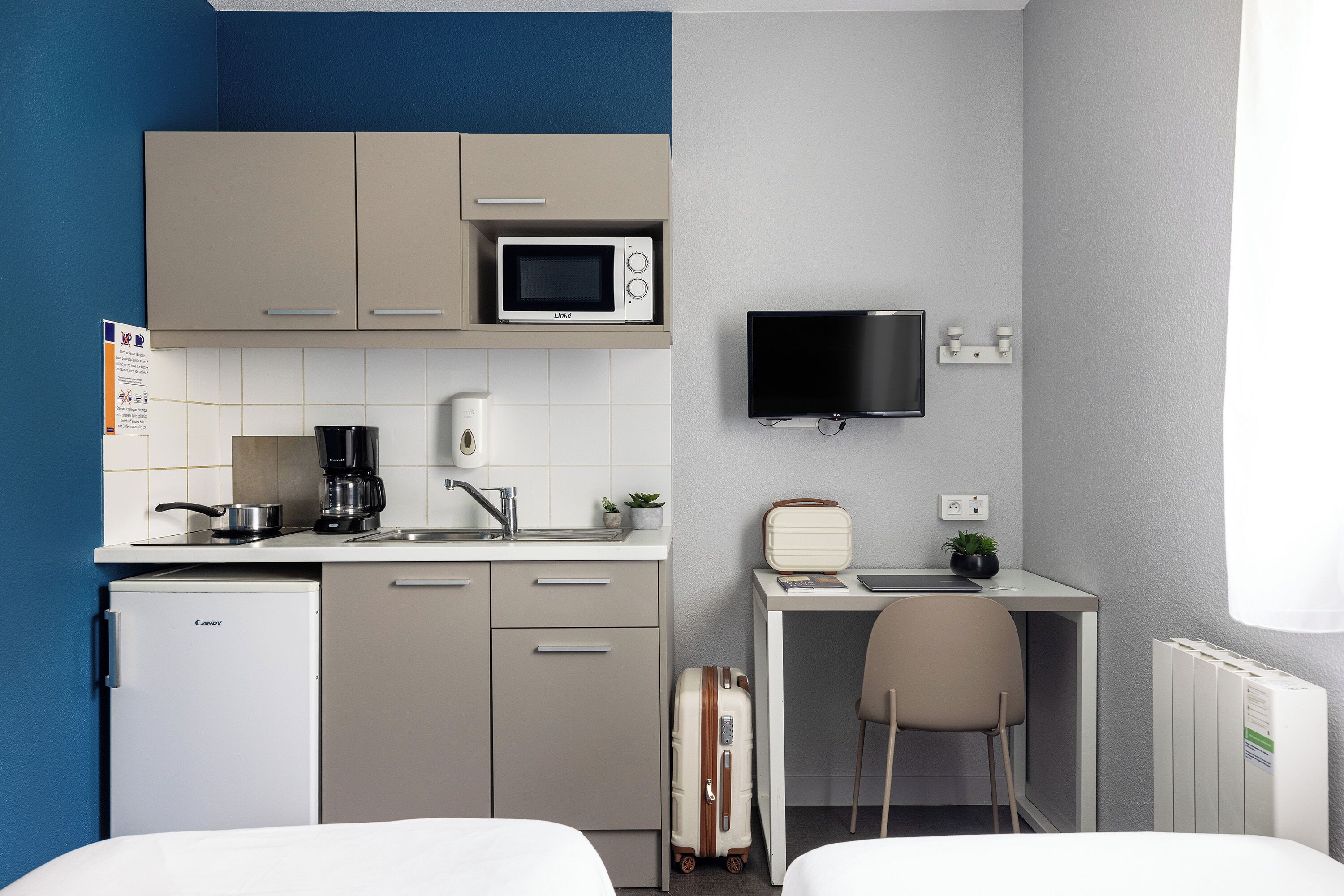 Habitación de hotel compacta con kitchenette, una cama, un escritorio con silla, un televisor y una maleta.
