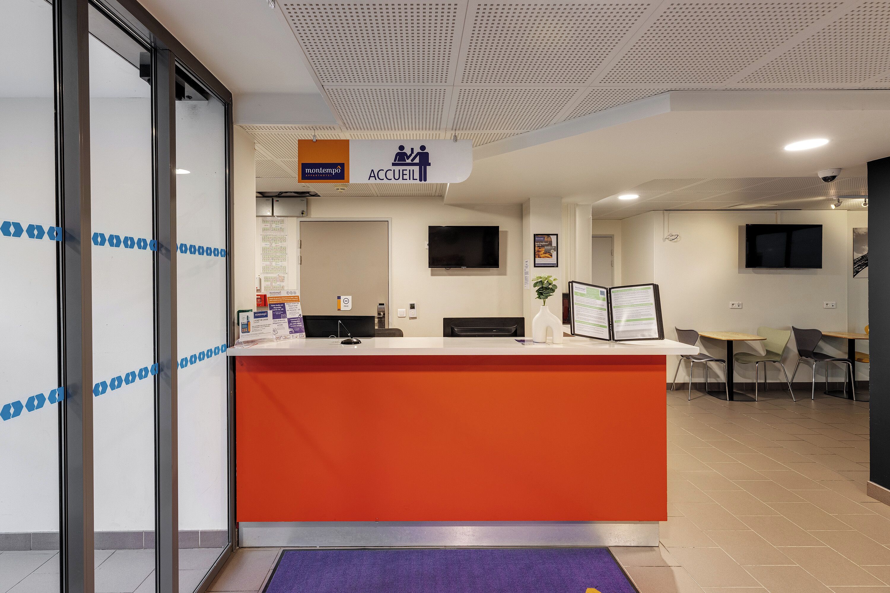 A reception desk with a sign indicating 'Accueil' and a seating area with tables and chairs.
