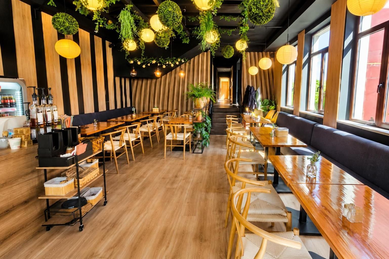 A restaurant interior with wooden tables and chairs, hanging plants, and a bar area with bottles.