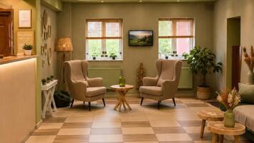 A hotel lobby with a checkered floor, two armchairs, a round table, a reception desk, and a television.