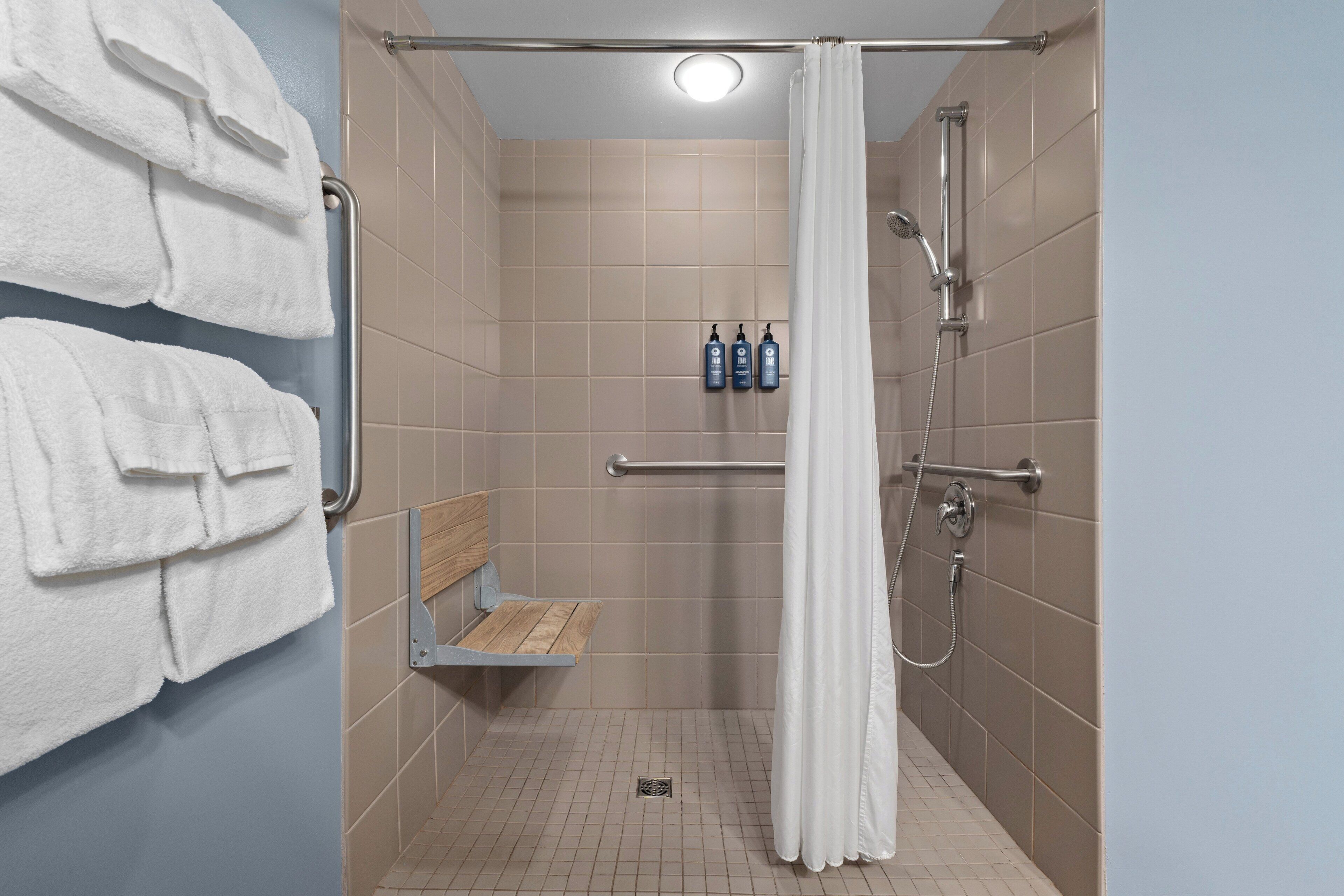 A shower area with a white shower curtain, a bench, and two bottles of shampoo.