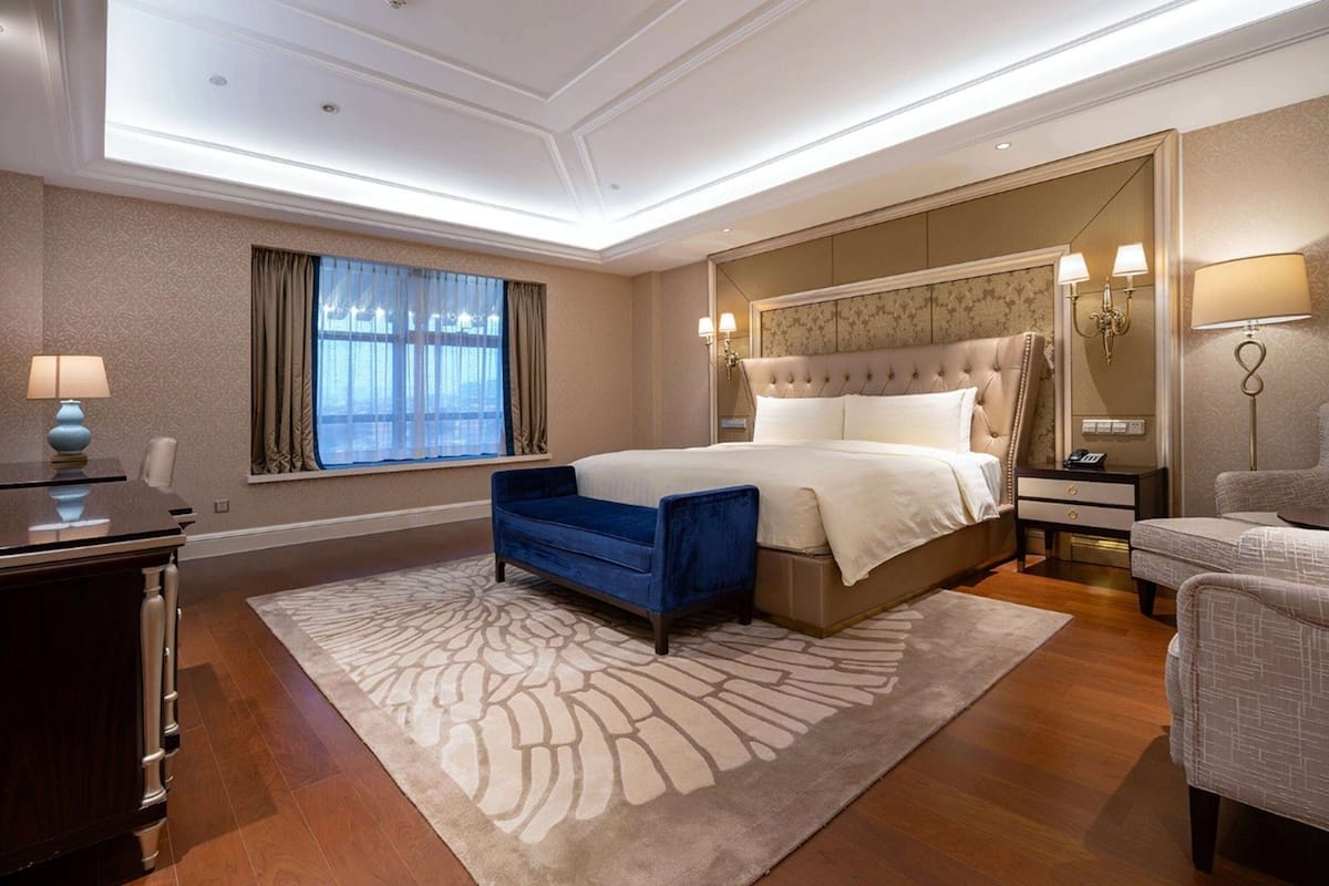 Un dormitorio amplio con una cama grande, una silla azul, un aparador de madera y una mesita de noche con lámpara.