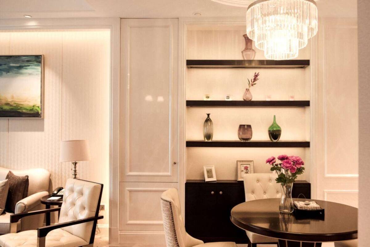 Una habitación de hotel moderna con decoración en tonos blancos, una araña, un sofá, una silla, una mesa redonda y una estantería con objetos decorativos.