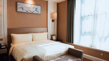 Una habitación de hotel con una cama grande, una mesita de noche con lámpara, una pequeña mesa con un marco de fotos y una ventana con cortinas.