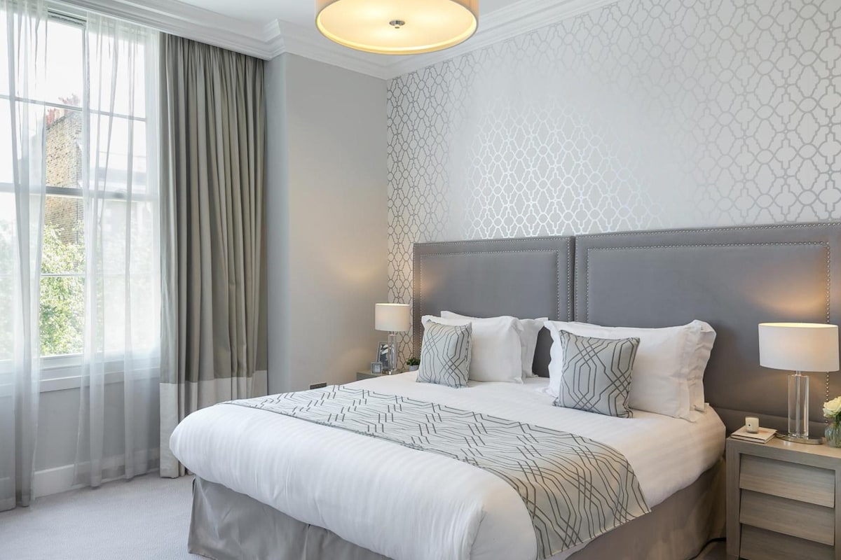 A hotel room with a large bed, two bedside tables, a headboard, and a window with curtains.