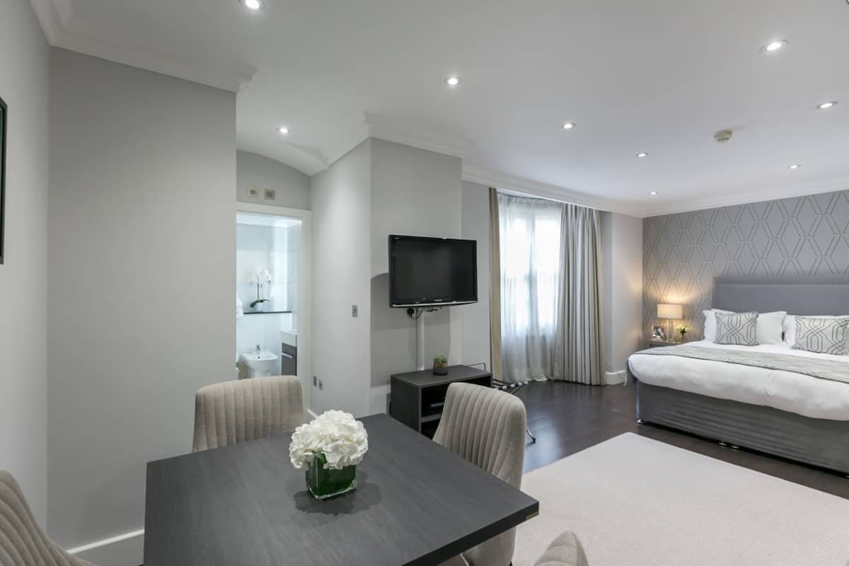A modern hotel room with a bed, a TV, a dining table with chairs, and a bathroom visible in the background.